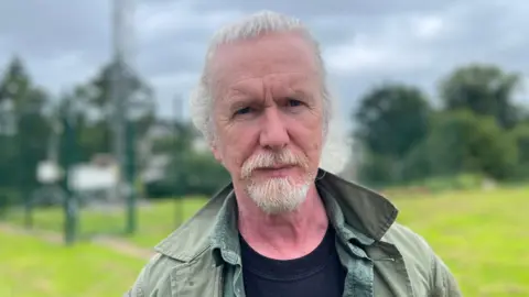 Shane Kelly, a man with white hair and beard, looks into the camera. He is wearing a green coat over a black t-shirt and a green shirt.