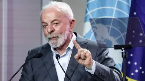 Shutterstock Brazilian President Luiz Inacio Lula da Silva speaks during the opening of the COP30 conference. He is wearing a grey suit and a white, open-necked shirt. He has white hair and a beard. He is holding up his left hand as he makes his speech. The UN and Brazil flags are behind him.
