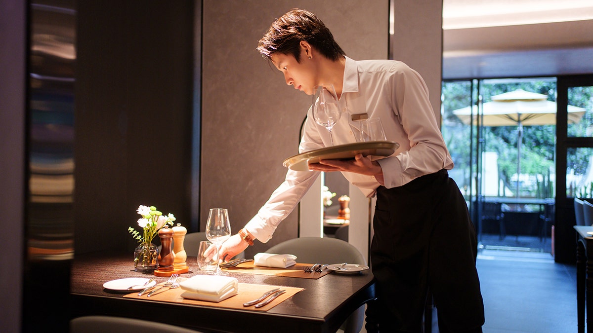 Waiter in uniform setting table at restaurant, placing glasses on table with flowers in the center.